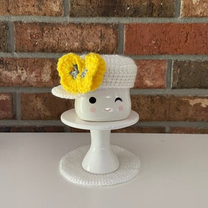 May include: White ceramic mug with a winking face, wearing a white crocheted hat with a yellow flower accent. The mug is sitting on a white crocheted coaster and a white pedestal.