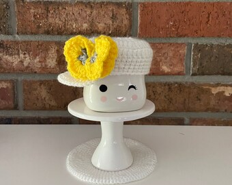May include: White ceramic mug with a winking face, wearing a white crocheted hat with a yellow flower accent. The mug is sitting on a white crocheted coaster and a white pedestal.