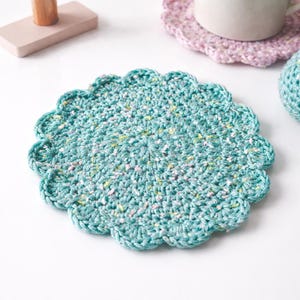 May include: A teal crocheted coaster with a scalloped edge. The coaster is round and has a speckled pattern of white, yellow, and pink. A pink crocheted coaster and a white mug are in the background.