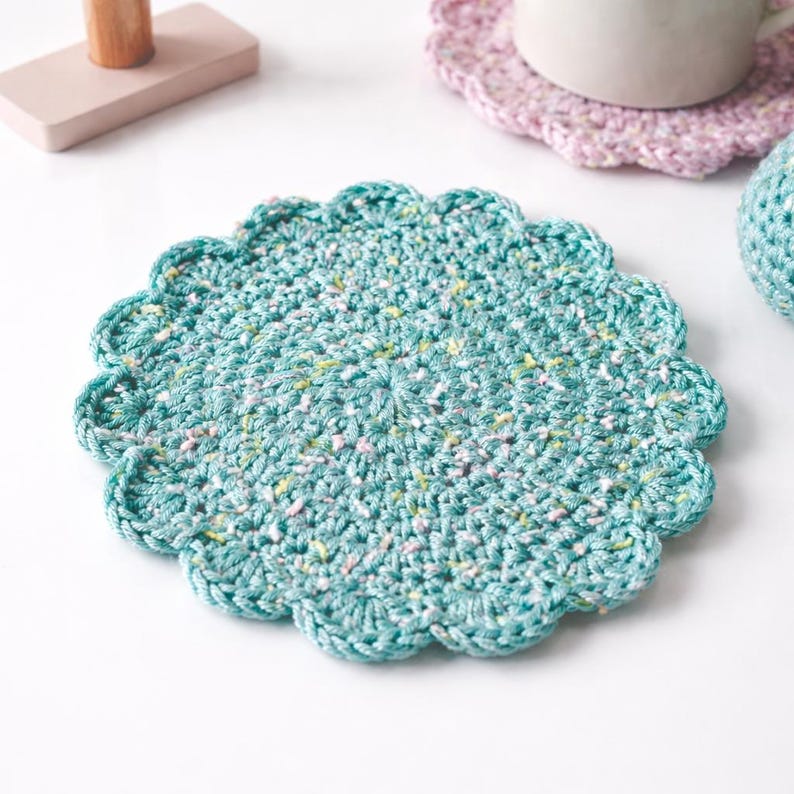 May include: A teal crocheted coaster with a scalloped edge. The coaster is round and has a speckled pattern of white, yellow, and pink. A pink crocheted coaster and a white mug are in the background.