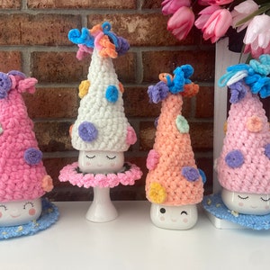 May include: Four crocheted mug cozies in different colors with smiling faces. The cozies are decorated with colorful polka dots and have a fluffy texture.