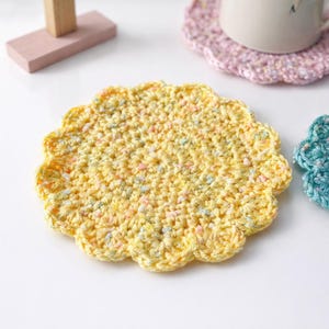 May include: A yellow, handmade crochet coaster with a scalloped edge. The coaster is made of yarn with flecks of blue, pink, and white. Other coasters in pink and teal are also visible.