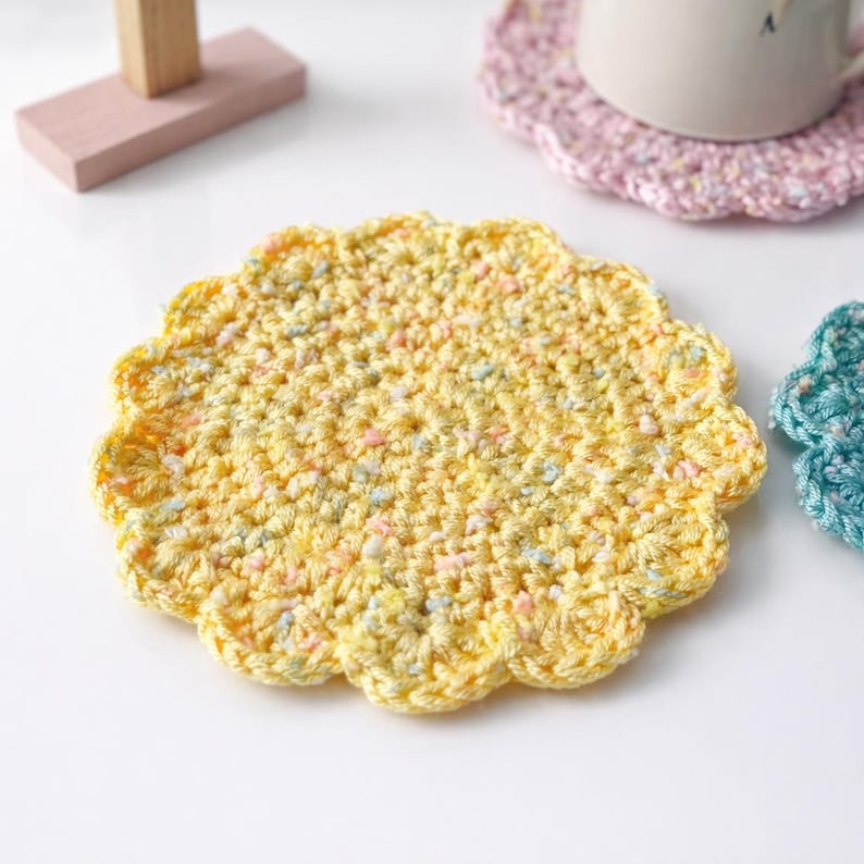 May include: A yellow, handmade crochet coaster with a scalloped edge. The coaster is made of yarn with flecks of blue, pink, and white. Other coasters in pink and teal are also visible.