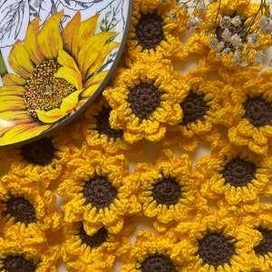 Pode incluir: Uma coleção de enfeites de girassol de crochê em amarelo e marrom. As flores estão dispostas em um padrão circular sobre uma superfície branca.
