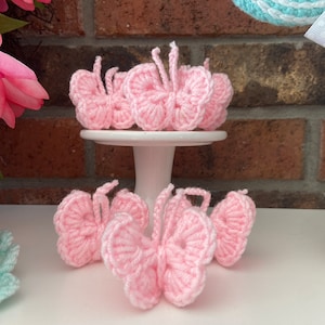May include: Several handmade, light pink crochet butterflies are displayed on a white pedestal and surface. The butterflies have detailed wing patterns and antennae. The background includes a brick wall and floral decorations.