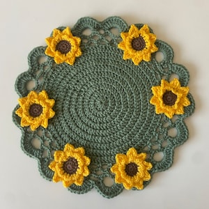 May include: Handmade crochet doily in a circular shape, featuring a dark green center and a scalloped edge. Adorned with seven yellow and brown sunflower appliqués, this decorative piece adds a touch of nature-inspired charm to any space.