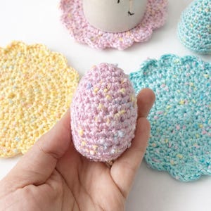 May include: A hand holding a pink crocheted egg with colorful flecks. Surrounding the egg are crocheted coasters in yellow, pink, and teal, and a mug with a floral design. The coasters have a scalloped edge.