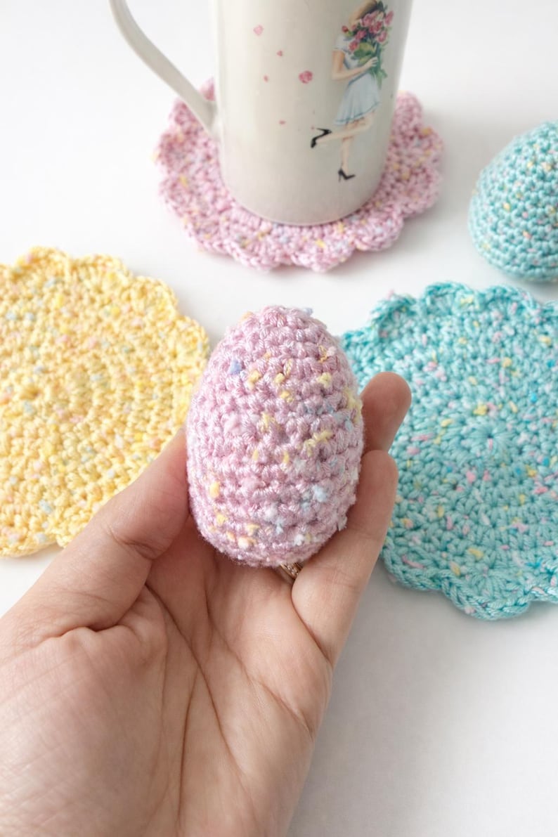 May include: A hand holding a pink crocheted egg with colorful flecks. Surrounding the egg are crocheted coasters in yellow, pink, and teal, and a mug with a floral design. The coasters have a scalloped edge.