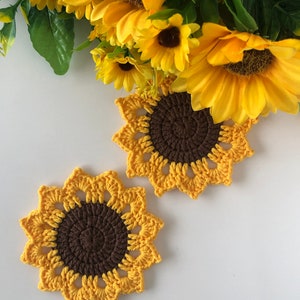 May include: Two crocheted sunflower coasters in yellow and brown. The coasters are on a white surface.