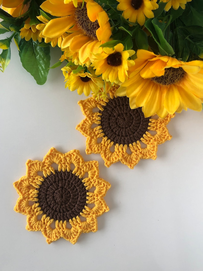 May include: Two crocheted sunflower coasters in yellow and brown. The coasters are on a white surface.