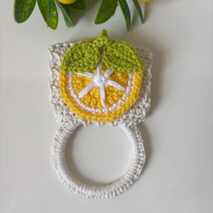 Puede incluir: Un soporte para toallas de ganchillo con una base cuadrada de color crema, decorado con un motivo de rodaja de lim&oacute;n en amarillo y blanco y dos hojas verdes. Incluye un anillo de ganchillo blanco en la parte inferior para colgar pa&ntilde;os.
