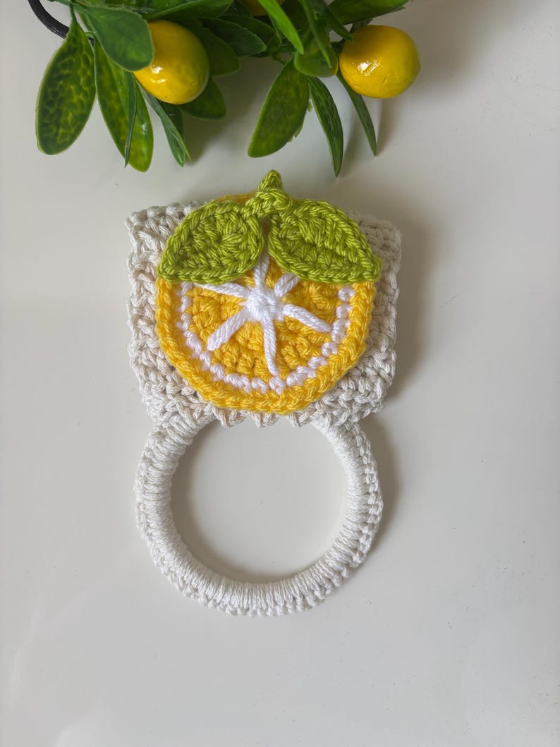 Puede incluir: Un soporte para toallas de ganchillo con una base cuadrada de color crema, decorado con un motivo de rodaja de lim&oacute;n en amarillo y blanco y dos hojas verdes. Incluye un anillo de ganchillo blanco en la parte inferior para colgar pa&ntilde;os.
