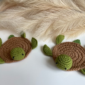 May include: Two crocheted turtle coasters in brown and green. The turtles have small black eyes and are lying on a white surface.
