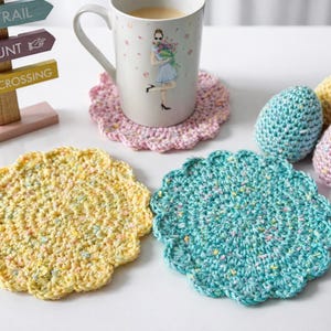 May include: A collection of handmade Easter decorations. Includes a mug with a floral design, crocheted coasters in yellow and teal, and three crocheted Easter eggs in blue, yellow, and pink. A wooden sign points to a bunny trail.