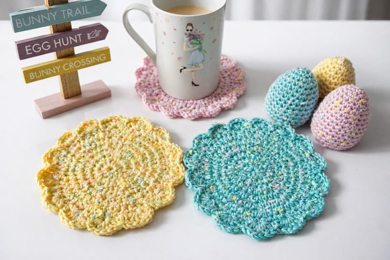 May include: A collection of handmade Easter decorations. Includes a mug with a floral design, crocheted coasters in yellow and teal, and three crocheted Easter eggs in blue, yellow, and pink. A wooden sign points to a bunny trail.