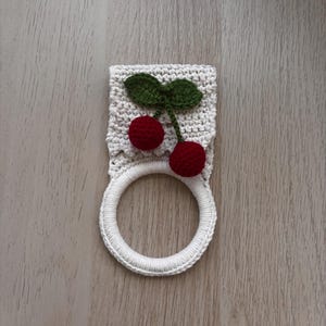 Puede incluir: Un soporte para toallas de ganchillo color crema con una aplicaci&oacute;n decorativa de dos cerezas de color rojo oscuro con hojas verdes. La parte superior es una pieza rectangular tejida y la inferior es un anillo circular de hilo blanco.