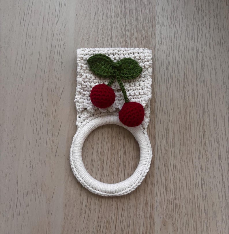 Puede incluir: Un soporte para toallas de ganchillo color crema con una aplicaci&oacute;n decorativa de dos cerezas de color rojo oscuro con hojas verdes. La parte superior es una pieza rectangular tejida y la inferior es un anillo circular de hilo blanco.