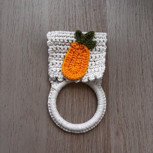 Puede incluir: Un soporte para toallas de ganchillo con una secci&oacute;n superior rectangular de color gris claro y patr&oacute;n de punto texturizado. Una aplicaci&oacute;n de zanahoria naranja con hojas verde oscuro est&aacute; centrada en la parte gris, unida a una base de anillo blanco.