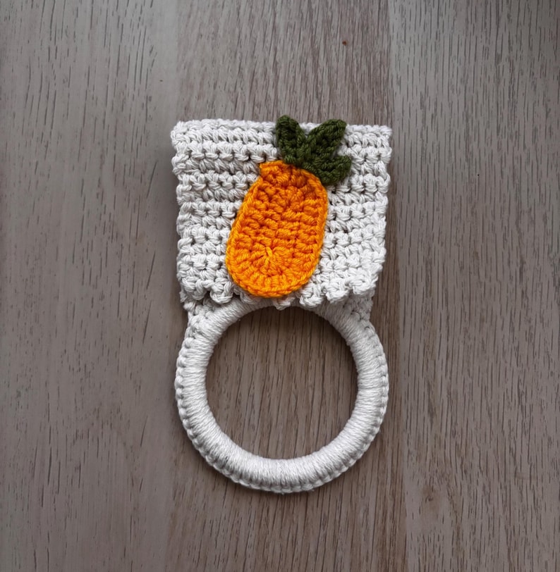 Puede incluir: Un soporte para toallas de ganchillo con una secci&oacute;n superior rectangular de color gris claro y patr&oacute;n de punto texturizado. Una aplicaci&oacute;n de zanahoria naranja con hojas verde oscuro est&aacute; centrada en la parte gris, unida a una base de anillo blanco.