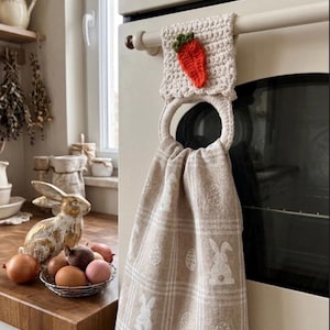 Puede incluir: Un pa&ntilde;o de cocina beige con un patr&oacute;n de cuadros blanco que presenta motivos de conejos y huevos, suspendido de un anillo de ganchillo color crema. La parte superior est&aacute; adornada con una peque&ntilde;a zanahoria naranja texturizada.