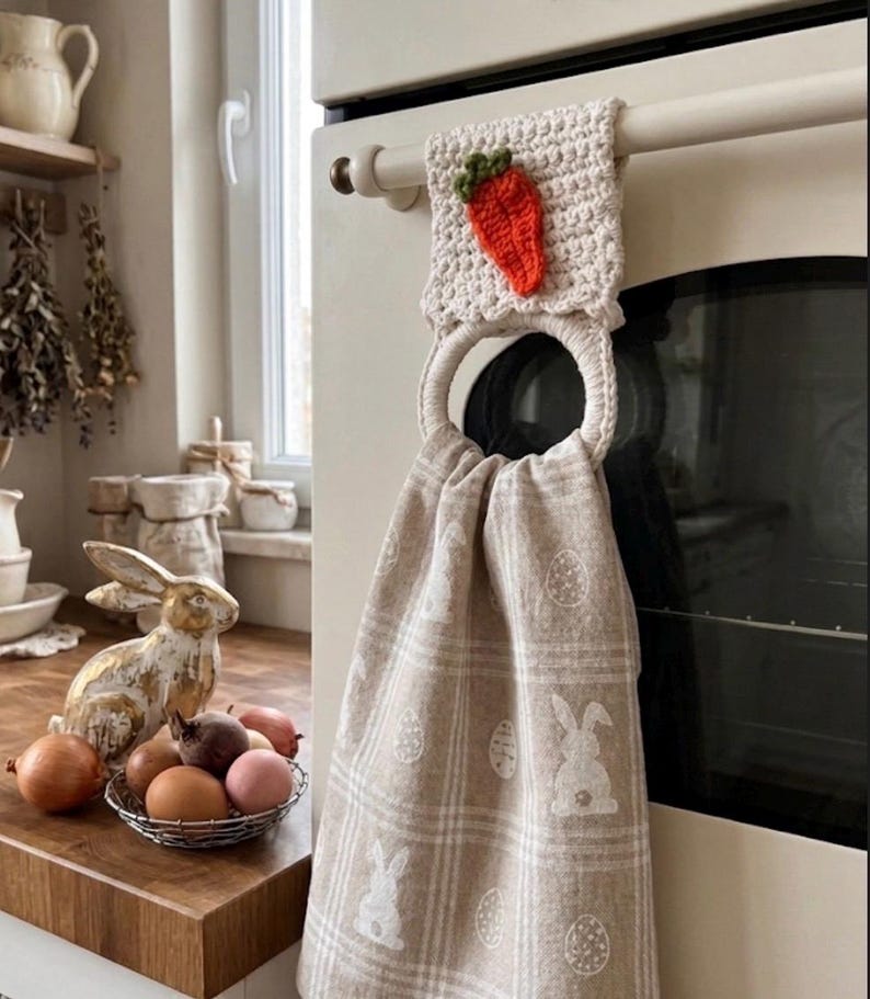 Puede incluir: Un pa&ntilde;o de cocina beige con un patr&oacute;n de cuadros blanco que presenta motivos de conejos y huevos, suspendido de un anillo de ganchillo color crema. La parte superior est&aacute; adornada con una peque&ntilde;a zanahoria naranja texturizada.