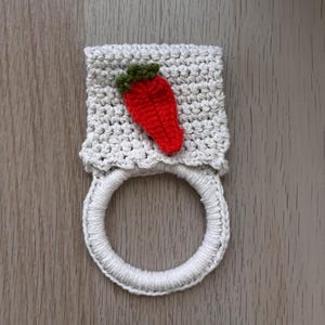 Puede incluir: Un toallero de ganchillo color crema con una secci&oacute;n superior rectangular texturizada adornada con un peque&ntilde;o aplique en forma de fresa roja con hojas verdes, unido a un anillo circular envuelto en hilo color crema a juego.