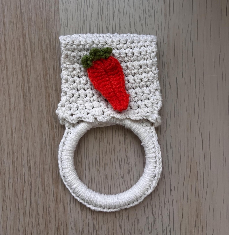 Puede incluir: Un toallero de ganchillo color crema con una secci&oacute;n superior rectangular texturizada adornada con un peque&ntilde;o aplique en forma de fresa roja con hojas verdes, unido a un anillo circular envuelto en hilo color crema a juego.