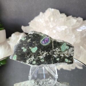 May include: A green and black stone with a purple and pink center. The stone is on a clear stand and is reflected in a mirror.