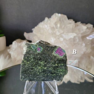 May include: A green and black stone with red and pink crystals embedded in it. The stone is on a clear plastic stand. The stone is labeled "B".