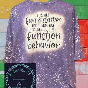 May include: Purple t-shirt with white bleach splatters and the text "IT'S ALL fun & games UNTIL SOMEONE FIGURES OUT THE function OF YOUR behavior".