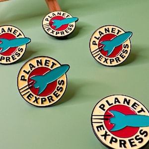 May include: A collection of Planet Express enamel pins, featuring the logo from Futurama. The pins are circular with a red background, a teal rocket, and the words "Planet Express". A small easel displays a matching sign.