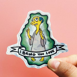 Könnte beinhalten: Eine gelbe Comicfigur mit blauen Haaren und großen Augen, die eine weiße Robe trägt, mit dem Text "I bring you love" auf einem Banner darunter. Der Aufkleber hat eine holografische Oberfläche.