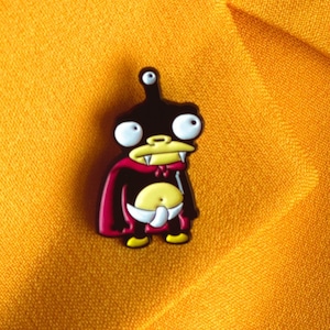 Peut inclure: Une épingle en émail noire et jaune représentant un personnage de dessin animé avec une cape rouge. Le personnage a de grands yeux, une petite bouche avec des crocs et une seule antenne sur la tête.
