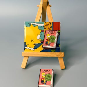 May include: A yellow and pink enamel pin featuring Bart Simpson sitting on a book with the text "I didn't do it! The Bart Simpson Story".