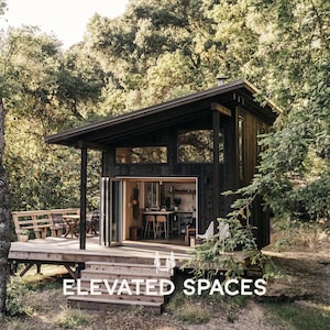 Op de afbeelding: Een donker gebeitste hut met een schuin dak en grote ramen, genesteld tussen bomen. De hut heeft een dek met een zithoek en de tekst "ELEVATED SPACES" onderaan. De open deuren onthullen een keuken en een eethoek.