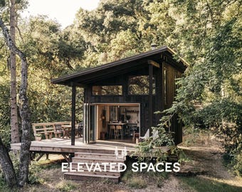 May include: A dark-stained cabin with a sloped roof and large windows, set amongst trees. The cabin has a deck with a seating area and the text "ELEVATED SPACES" at the bottom. The open doors reveal a kitchen and dining area.