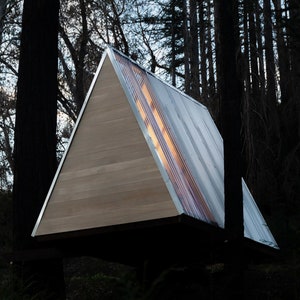 Puede incluir: Una peque&ntilde;a caba&ntilde;a de madera de forma triangular con un techo de metal. La caba&ntilde;a tiene una gran ventana en la parte delantera y el techo est&aacute; hecho de chapa corrugada. La caba&ntilde;a est&aacute; rodeada de &aacute;rboles.