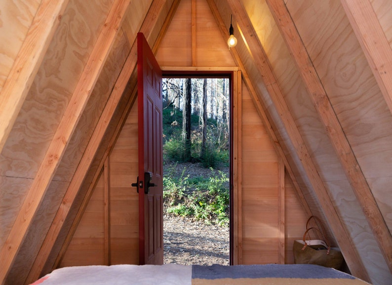 Pu&ograve; includere: Una capanna di legno a forma di A con una porta rossa aperta che rivela una vista di un bosco. La capanna ha un soffitto e pareti in legno, e un letto &egrave; visibile in primo piano.