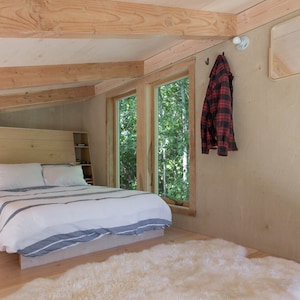 May include: A bedroom with a wooden bed frame, white bedding, and a white sheepskin rug. There are two windows with views of trees outside. A red and black plaid shirt is hanging on a hook on the wall.