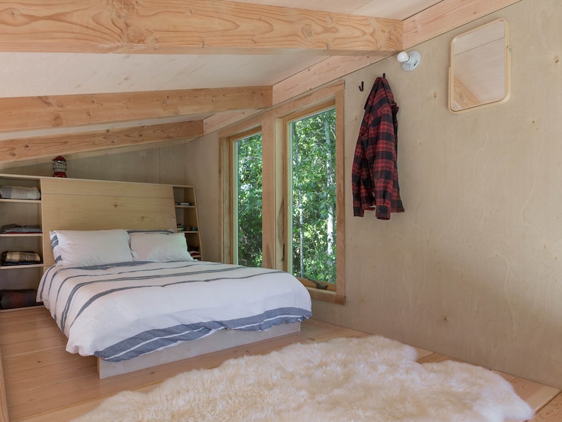 May include: A bedroom with a wooden bed frame, white bedding, and a white sheepskin rug. There are two windows with views of trees outside. A red and black plaid shirt is hanging on a hook on the wall.
