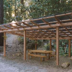 May include: A wooden outdoor dining set with a table and two benches under a covered pergola with string lights. The pergola is made of wood and has a corrugated metal roof.