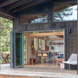 May include: A modern cabin with a wooden exterior and large windows. The cabin has a deck with a table and chairs, and a white chair on the deck. The interior of the cabin is visible through a set of folding glass doors, revealing a kitchen and living area.