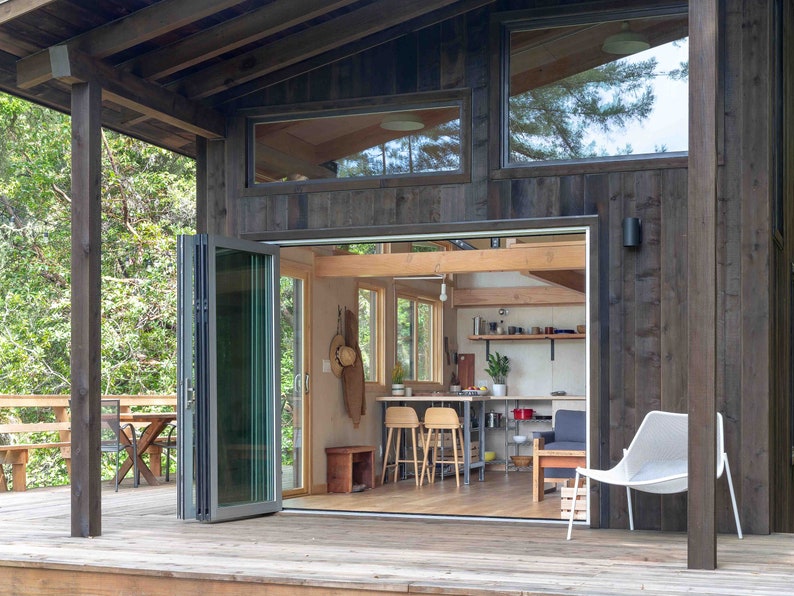 May include: A modern cabin with a wooden exterior and large windows. The cabin has a deck with a table and chairs, and a white chair on the deck. The interior of the cabin is visible through a set of folding glass doors, revealing a kitchen and living area.