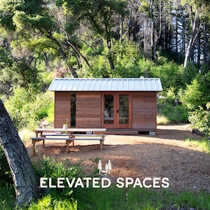 Puede incluir: Una cabaña rectangular de madera con techo de metal plateado, puertas dobles de cristal y una ventana vertical. Una mesa de picnic de madera se sitúa frente a la estructura. El texto ELEVATED SPACES aparece abajo con un icono de hamaca.