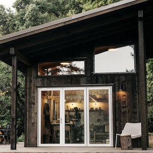 May include: A dark wood cabin with a large window and a set of glass doors. The cabin is surrounded by trees and has a small deck with a table and chairs.