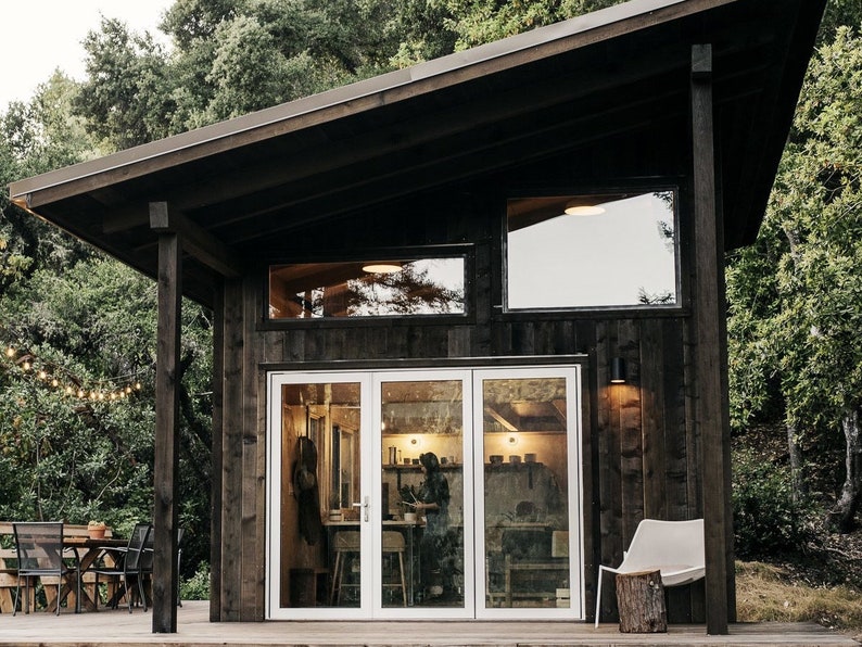 May include: A dark wood cabin with a large window and a set of glass doors. The cabin is surrounded by trees and has a small deck with a table and chairs.