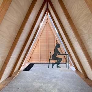 Puede incluir: Una persona se sienta en un escritorio en una habitaci&oacute;n triangular con paneles de madera y una ventana. La habitaci&oacute;n tiene una alfombra gris claro y una alfombra negra.