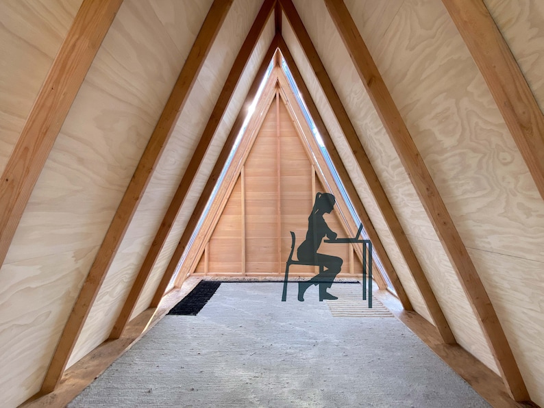 Puede incluir: Una persona se sienta en un escritorio en una habitaci&oacute;n triangular con paneles de madera y una ventana. La habitaci&oacute;n tiene una alfombra gris claro y una alfombra negra.