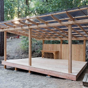 May include: A wooden outdoor structure with a corrugated plastic roof and a wooden deck. The structure has a wooden fence behind it and a wooden table and chairs on the deck.