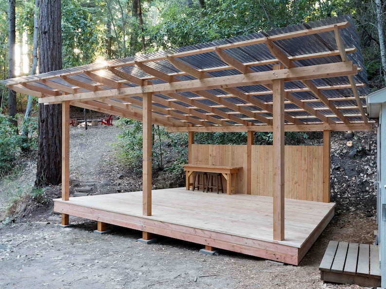 May include: A wooden outdoor structure with a corrugated plastic roof and a wooden deck. The structure has a wooden fence behind it and a wooden table and chairs on the deck.